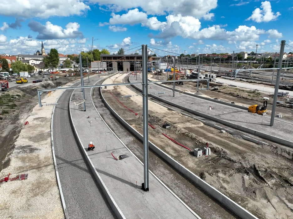 Chantier ferroviaire en construction avec voies courbes en béton, poteaux caténaires installés, ouvriers en équipement de sécurité, machines de terrassement jaunes, bâtiment en construction à l'arrière-plan, zone urbaine visible